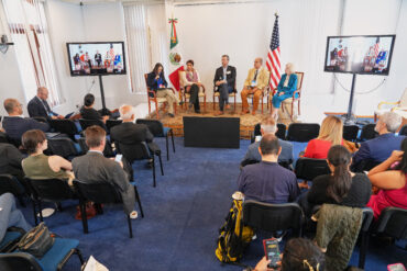 Wide view of a conference room with an audience seated and a panel of speakers on stage, flanked by the Mexican and U.S. flags, with two monitors displaying the live discussion.