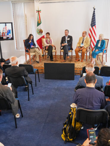 Wide view of a conference room with an audience seated and a panel of speakers on stage, flanked by the Mexican and U.S. flags, with two monitors displaying the live discussion.