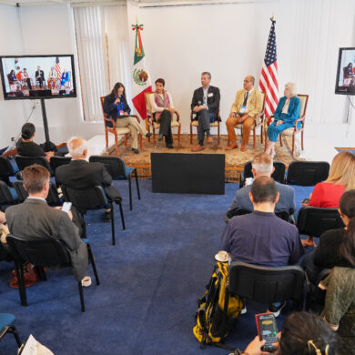 Wide view of a conference room with an audience seated and a panel of speakers on stage, flanked by the Mexican and U.S. flags, with two monitors displaying the live discussion.
